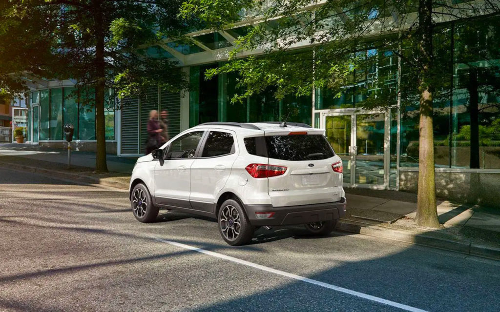 White SUV parked on urban street, surrounded by glass buildings and trees, showcasing modern city life.