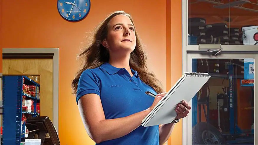 Woman in a blue polo with clipboard, standing in an auto repair shop, looking determined.