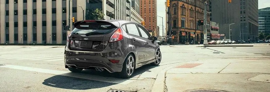 Compact car on urban street corner, surrounded by tall buildings and traffic lights.