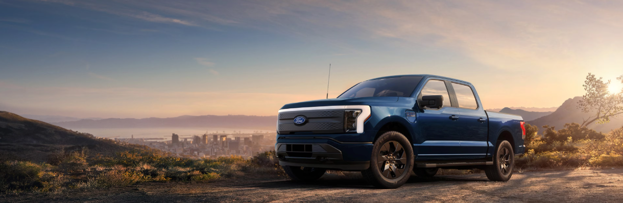 A blue 2025 Ford F-150 Lightning is parked on a dirt road overlooking a city skyline at sunset.